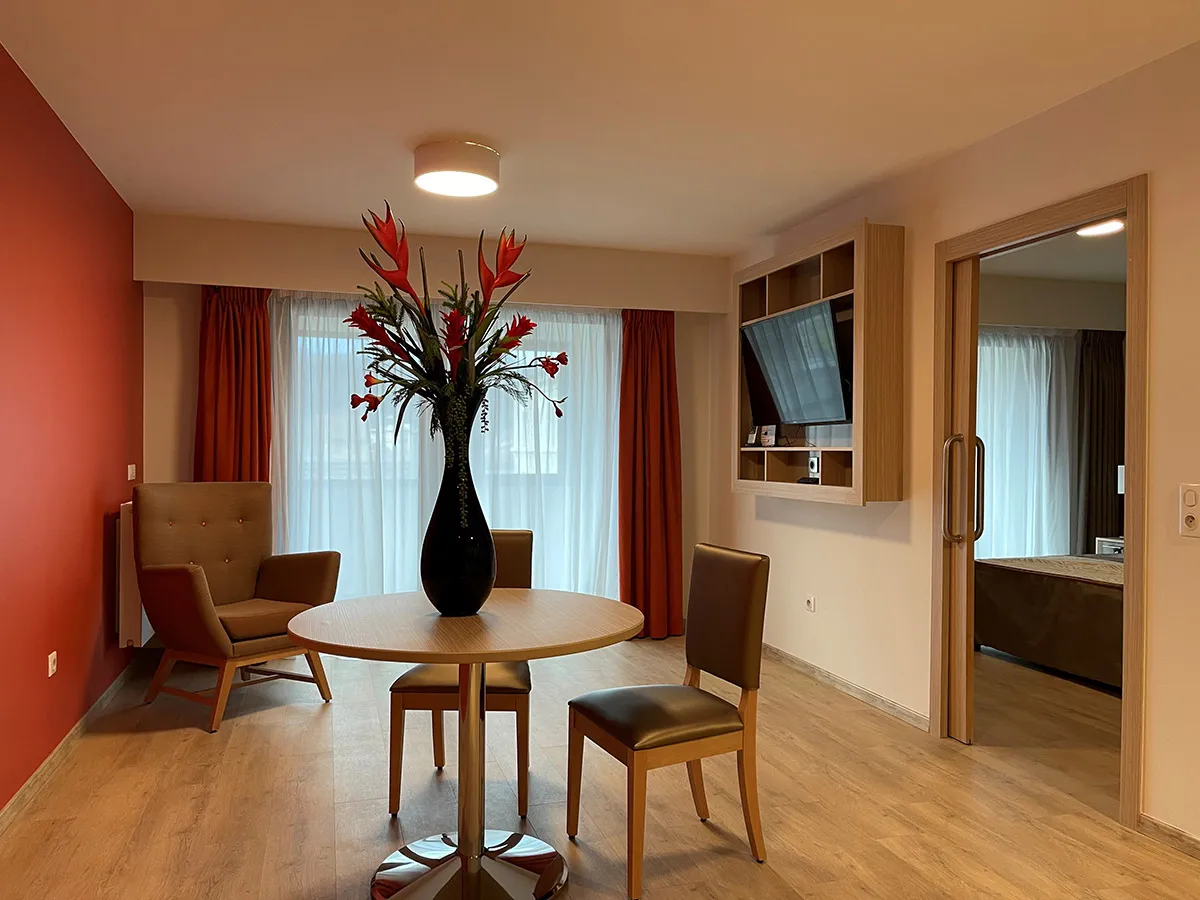 Living room of an apartment at the OBEO senior residence in Aix-les-Bains, with a red accent wall, round table with flowers, armchair and the bedroom visible through the sliding door