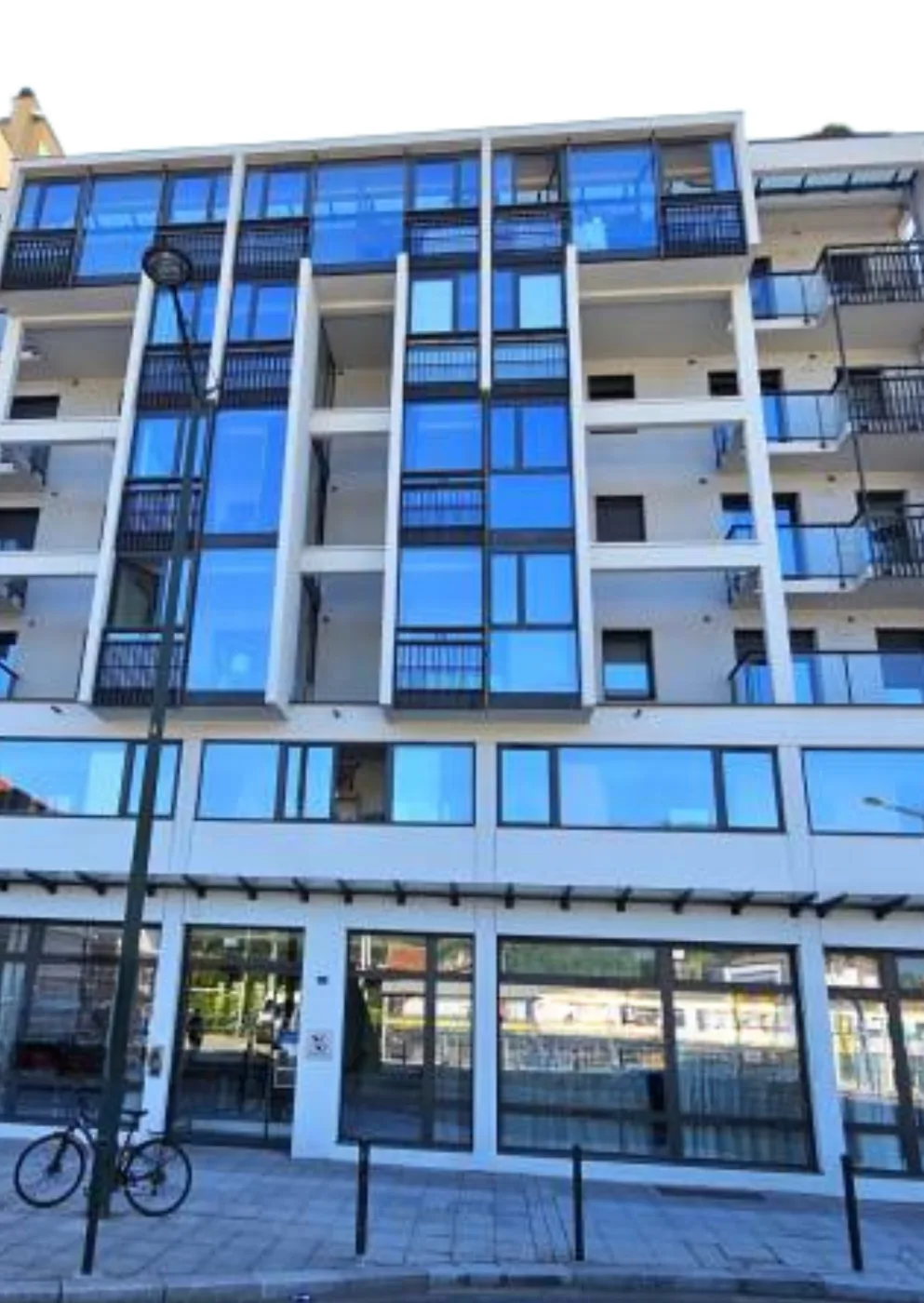 Street view of the OBEO Aix-les-Bains residence: six storeys of blue-tinted glass balconies, shops at ground level and a bicycle parked along the pavement