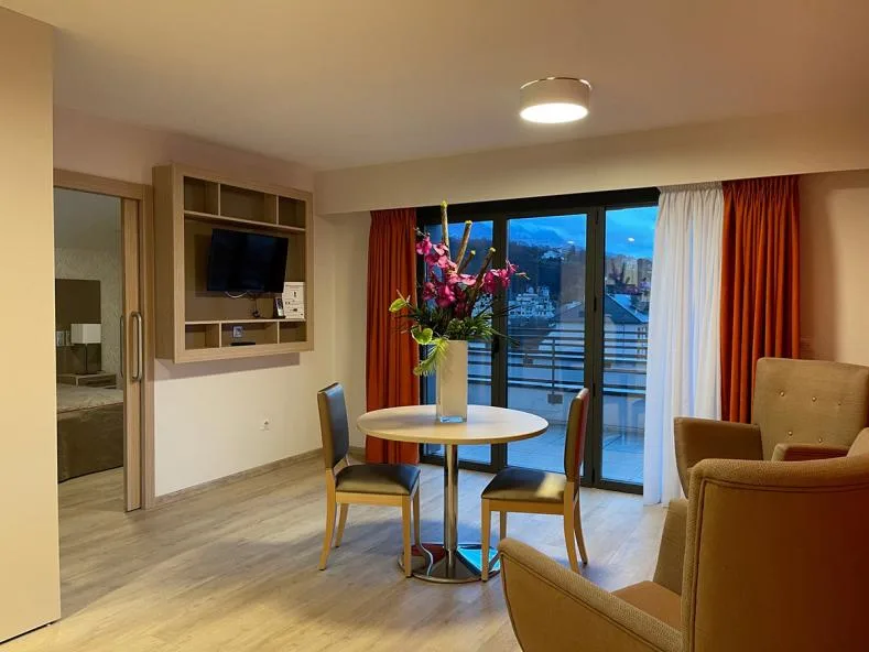 Living area of a show apartment in Aix-les-Bains: round table with flowers, armchairs, built-in television and a glazed door opening onto the balcony and the mountains