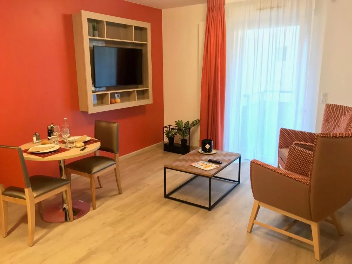 Séjour d’un appartement témoin de Chambéry : mur accent rouge, téléviseur dans une niche en bois, table de repas dressée et fauteuils près de la baie vitrée
