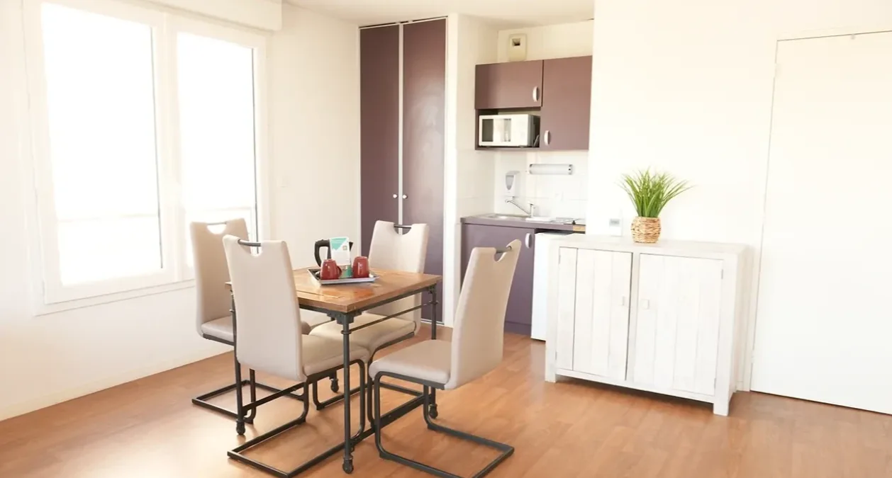 Dining corner of a show apartment in Nevers: wooden table and white chairs in front of the kitchenette and sideboard, below a large window