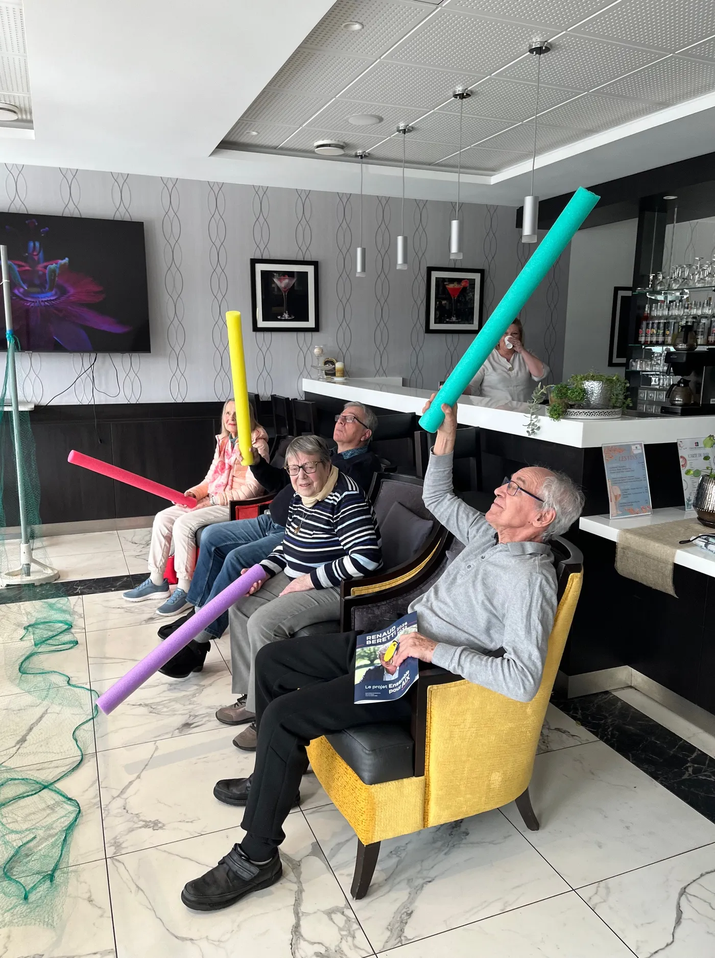 Deux résidents lèvent des bâtons en mousse colorés au-dessus de leur tête depuis leurs fauteuils