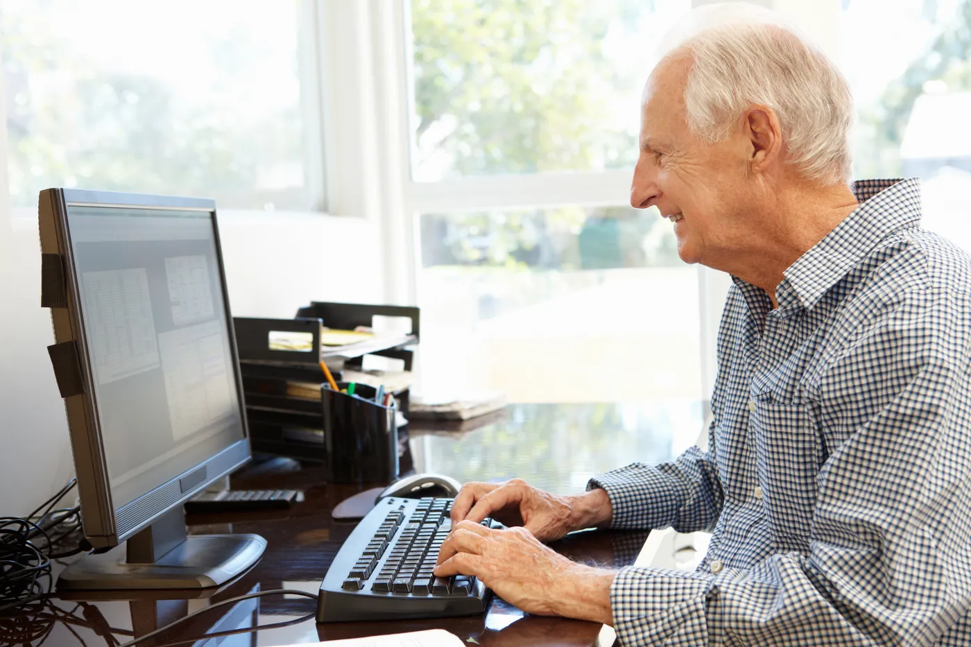 An older man at his desk by the window, typing on a keyboard in front of a large screen