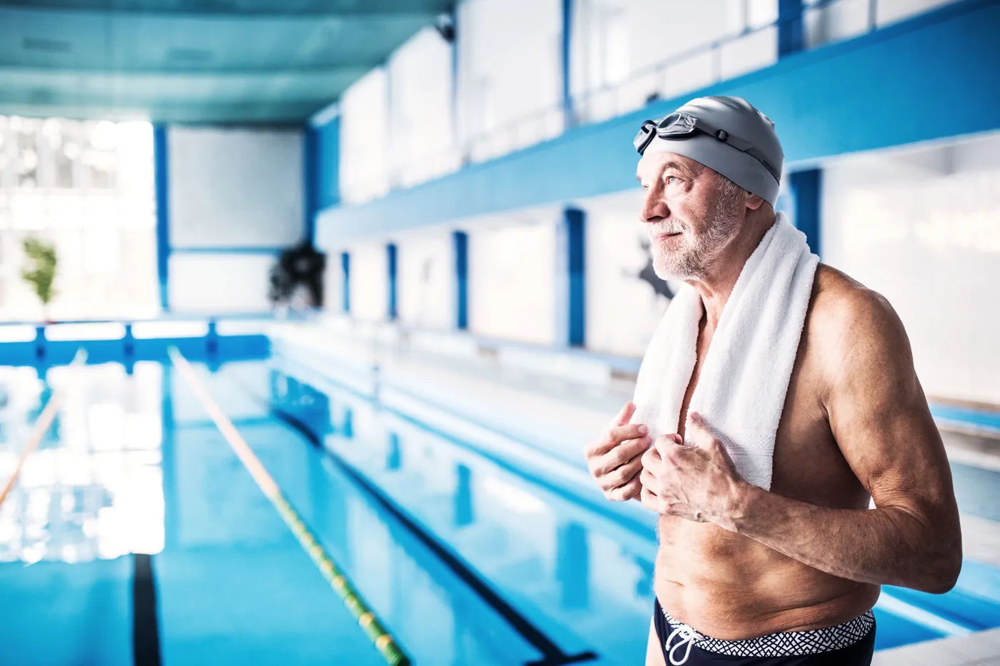 Un homme âgé en bonnet et maillot de bain, serviette sur les épaules, au bord d'un bassin de nage couvert