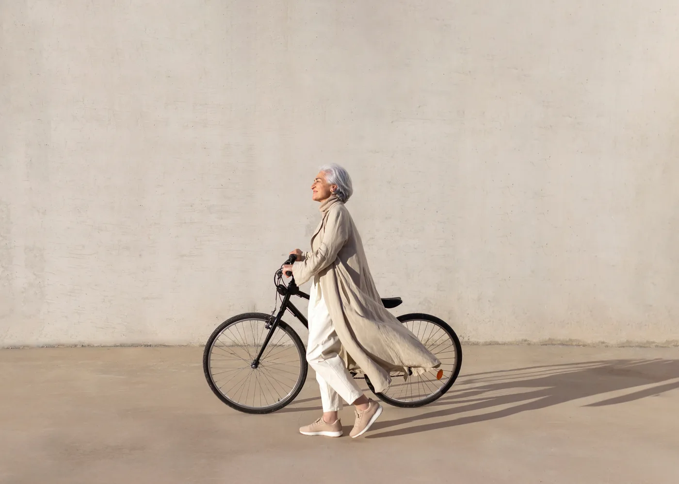 Une femme aux cheveux gris marche à côté de son vélo devant un mur clair ensoleillé