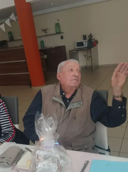 An elderly man raising his hand in greeting, a wrapped gift set down in front of him