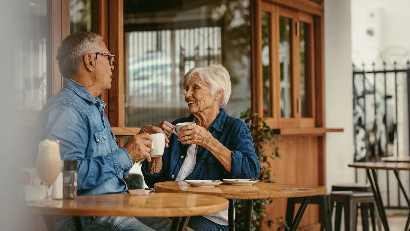 A senior couple sitting at a cafe table with cups of coffee, talking quietly