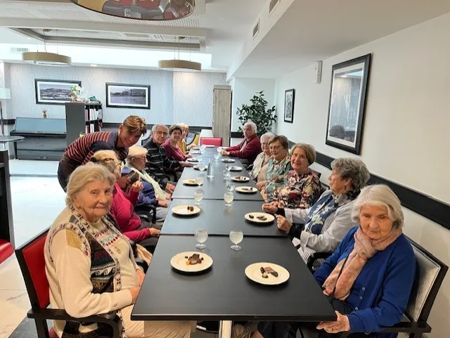 Residents seated along the long table, each with a plate of chocolates and a glass of water