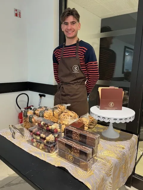 A young chocolatier in a striped apron standing behind his display of pastries and chocolates