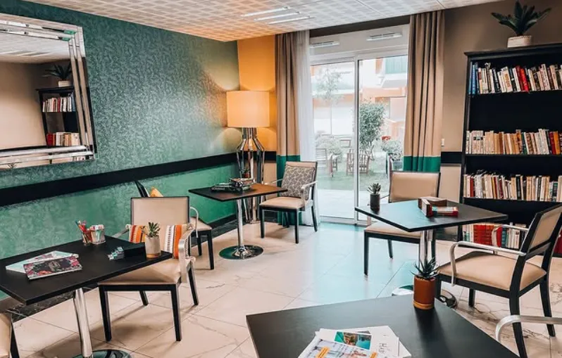 Un salon résidents aux murs lambrissés de vert, avec bibliothèque et fauteuils face à la terrasse