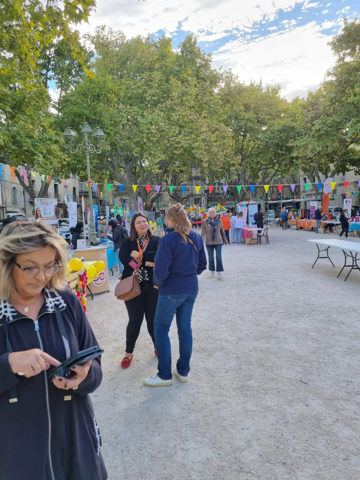 Une visiteuse consulte son téléphone au milieu de la place pendant la fête des aidants, les stands en arrière-plan