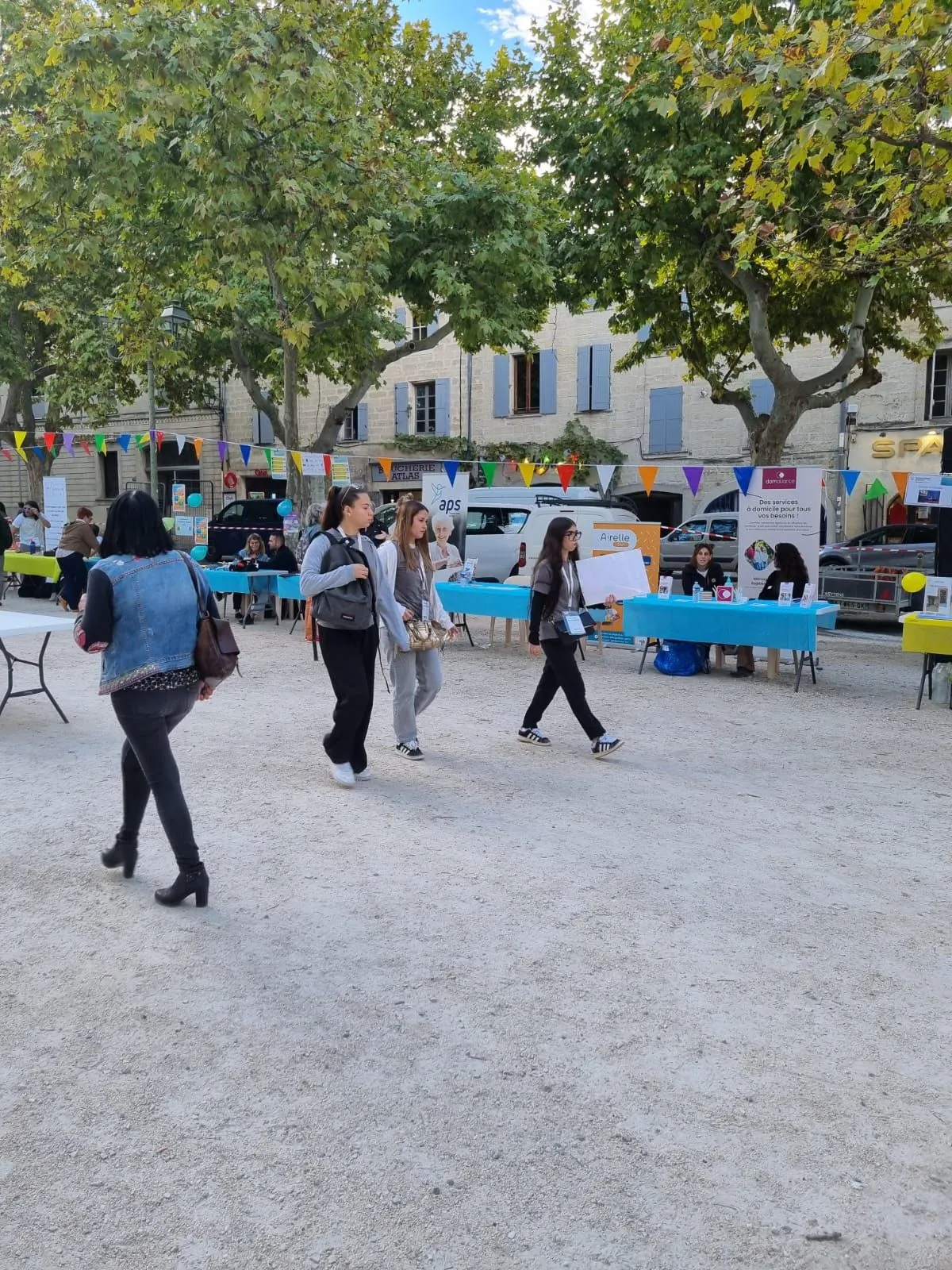Des passants traversent la place de village ombragée, des guirlandes de fanions tendues entre les platanes