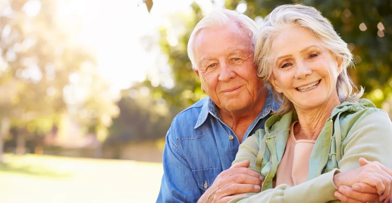 A senior couple leaning together and smiling, sunlit trees behind them