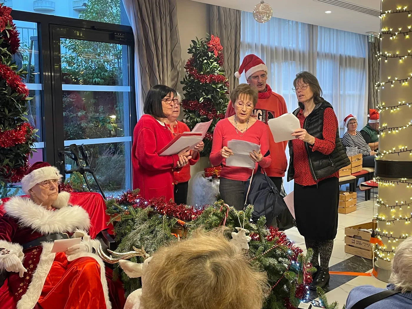 A choir in red singing carols beside Father Christmas in the decorated lounge