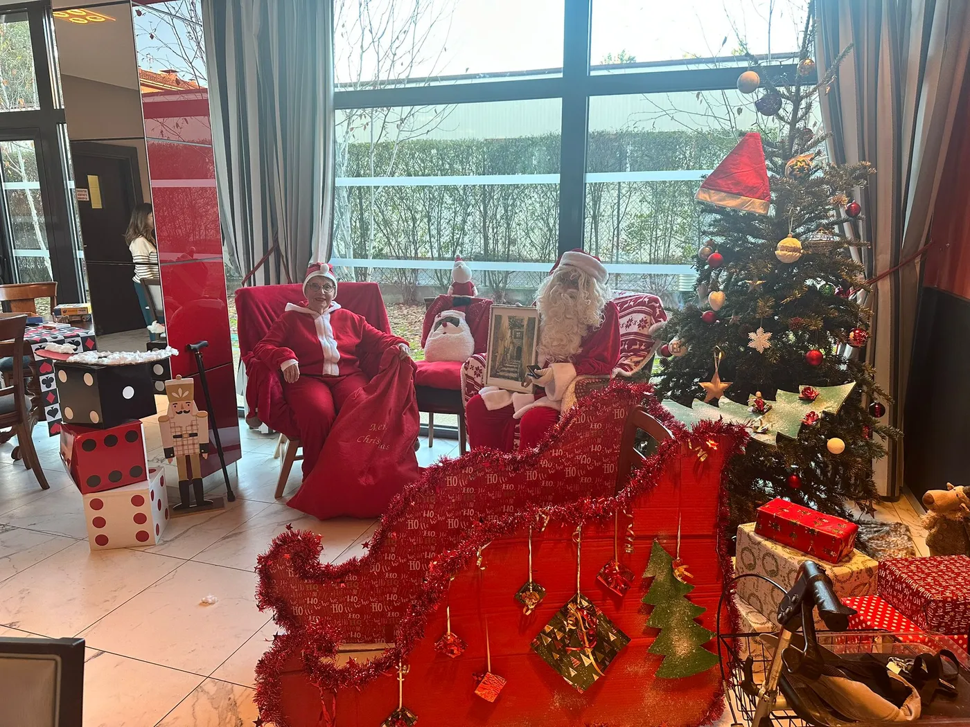 Un coin salon paré de rouge : un traîneau garni de guirlandes et de décorations, des cadeaux et des personnages en costume de Père Noël installés dans les fauteuils
