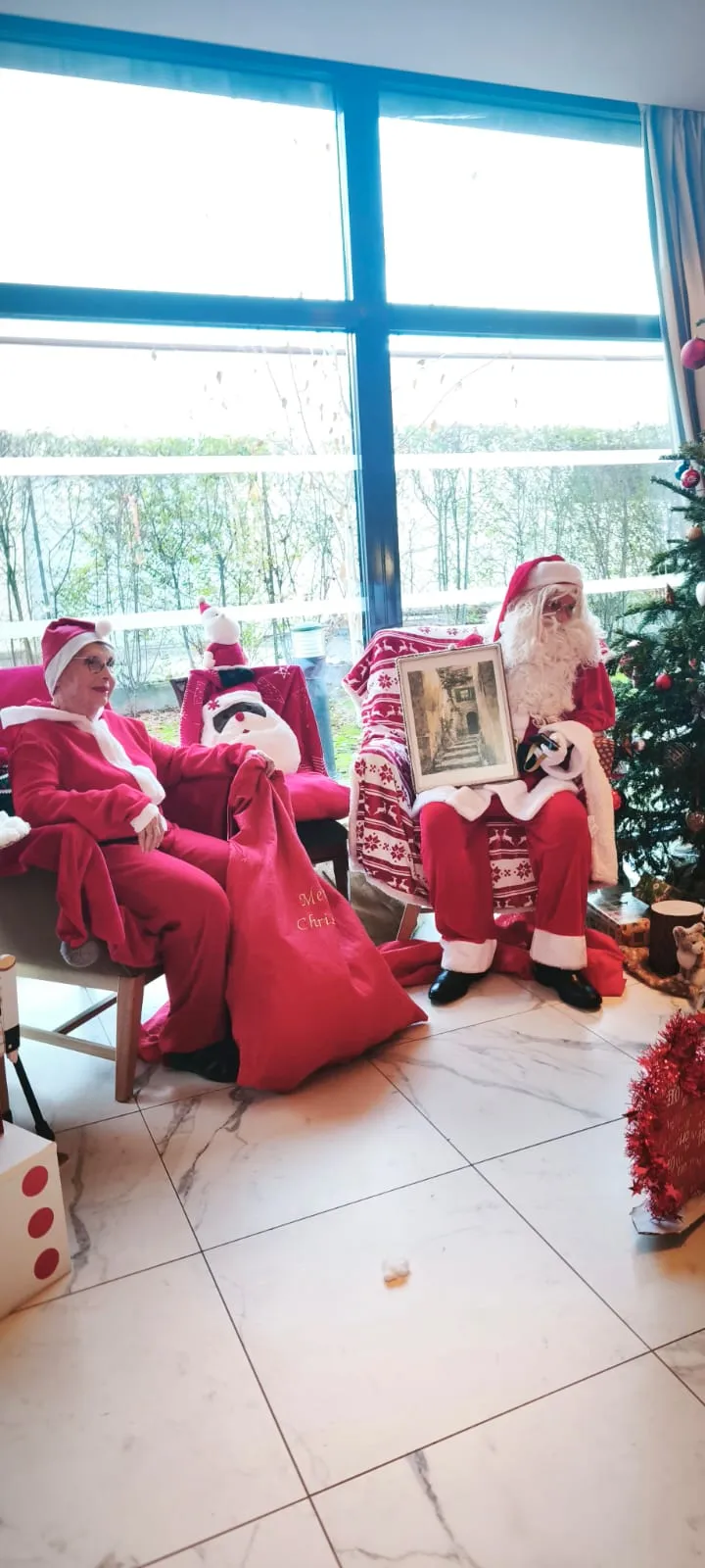 Deux personnages en costume rouge installés dans des fauteuils devant la baie vitrée, entourés de décorations de Noël