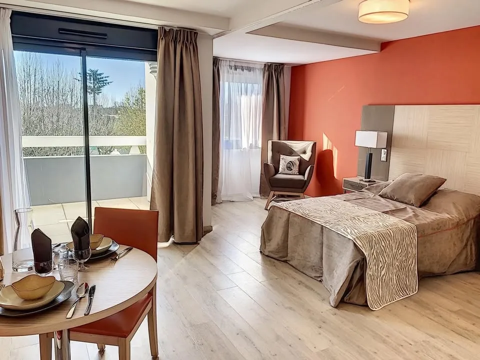 Un appartement meublé avec mur rouge, lit simple et table dressée près de la fenêtre