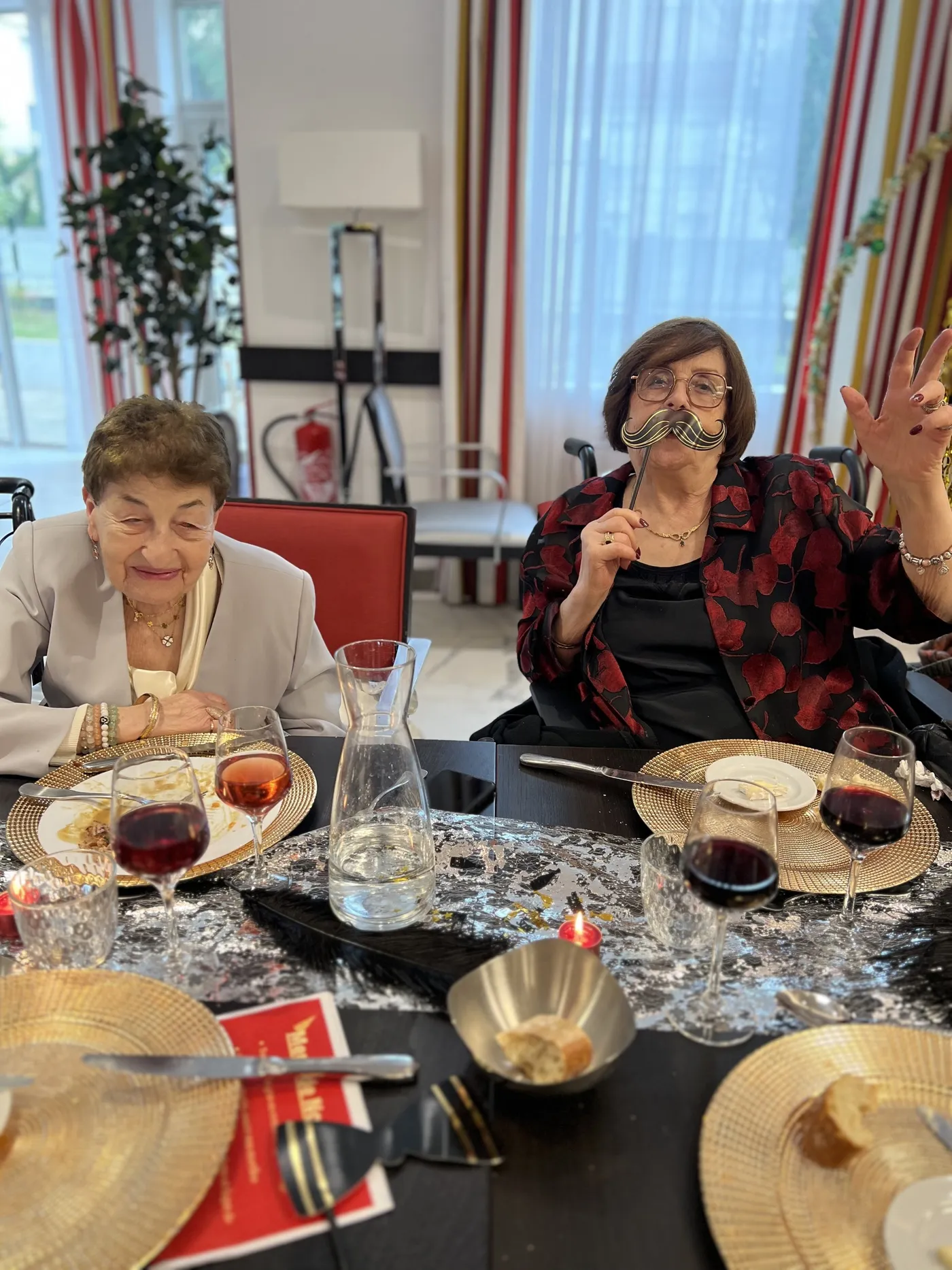 Two residents at the table, one waving, with gold chargers and glasses of wine