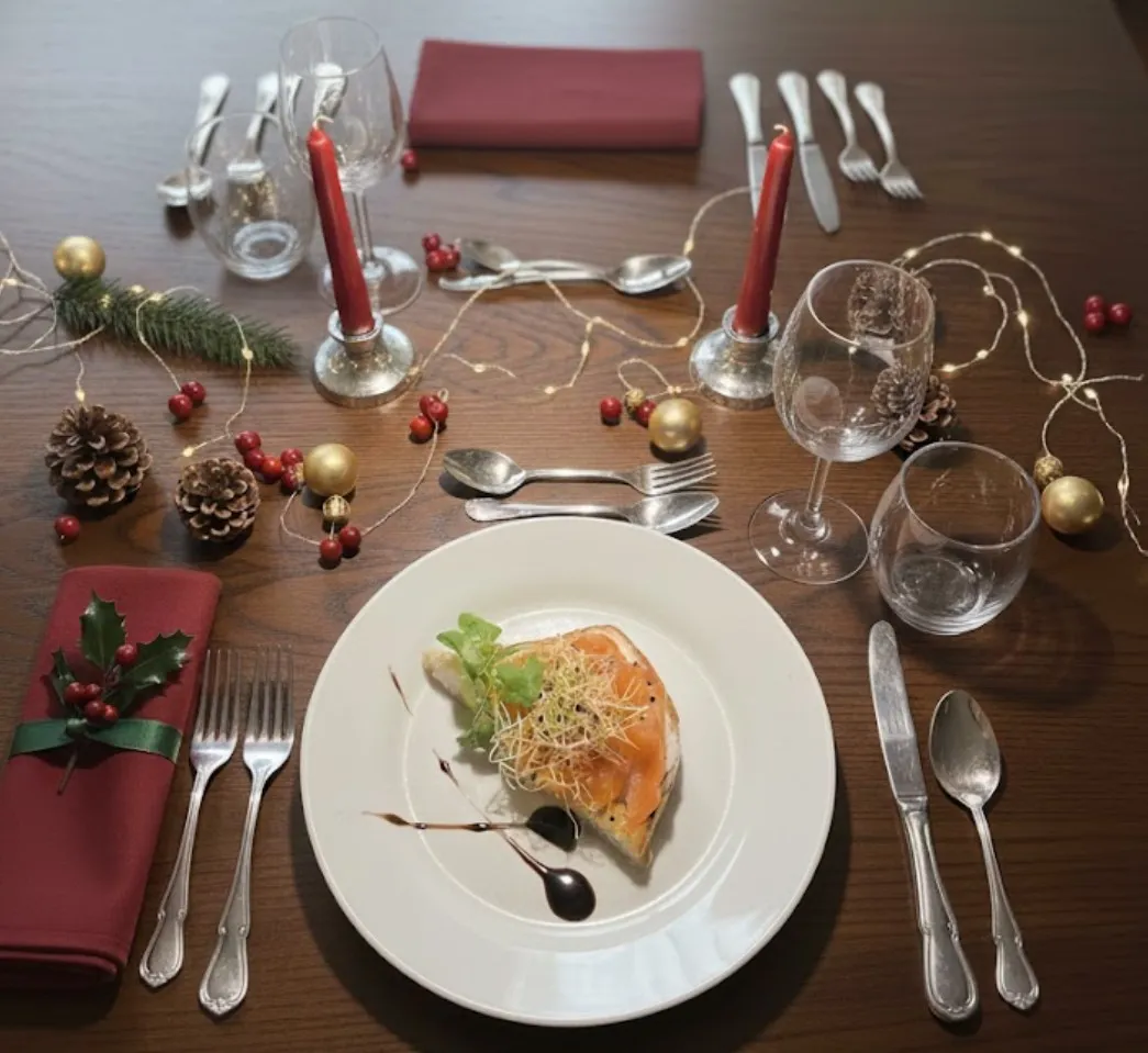 Une table de Noël dressée avec bougies rouges, pommes de pin, boules dorées et guirlande lumineuse, et une assiette de saumon fumé garni de jeunes pousses