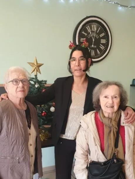 Une membre de l'équipe entoure de ses bras deux résidentes près du sapin de Noël