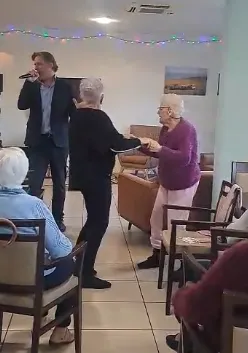 Le chanteur en costume tend la main à une résidente pendant que les autres suivent la scène depuis leurs fauteuils