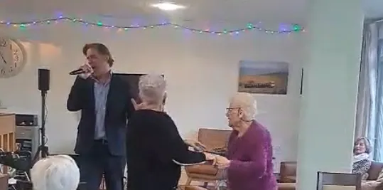 Micro en main, le chanteur interprète un titre près de l'enceinte, sous l'horloge du salon, entouré de deux résidentes