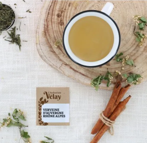 A cup of herbal tea seen from above on a wooden board, with cinnamon sticks and a packet of verbena