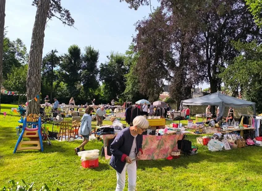 Flea market set up on the lawn of wooded grounds: stalls under a gazebo, clothes rails and tables of second-hand goods, visitors browsing in the sunshine.