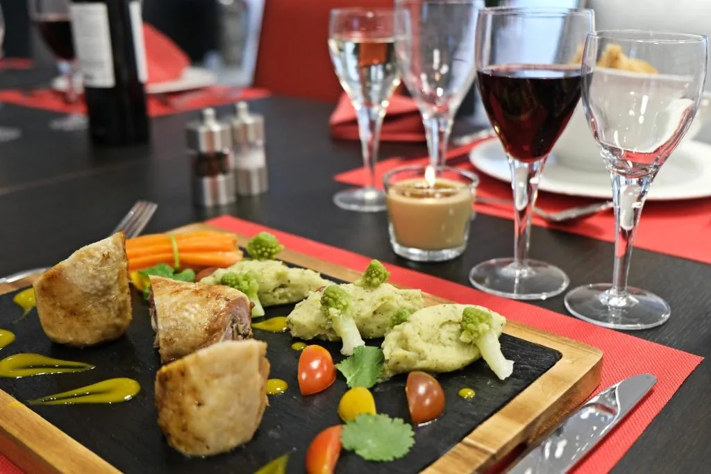 Plated dish: meat, carrots, broccoli and cherry tomatoes on slate, wine glasses in the background