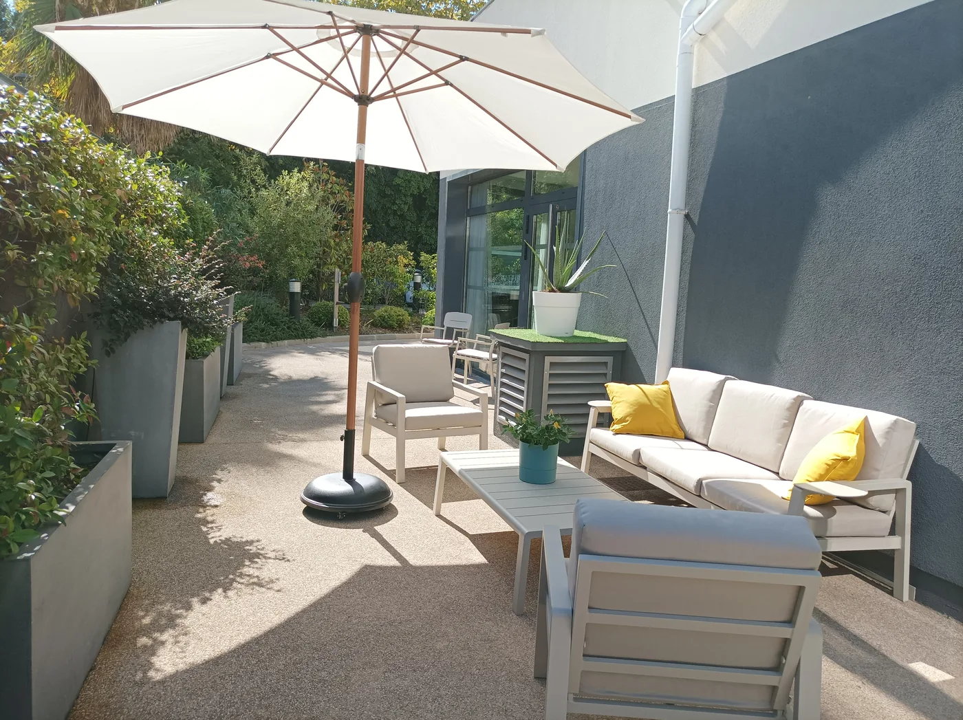 Une terrasse ombragée par un grand parasol, avec canapé et fauteuils installés entre des massifs plantés