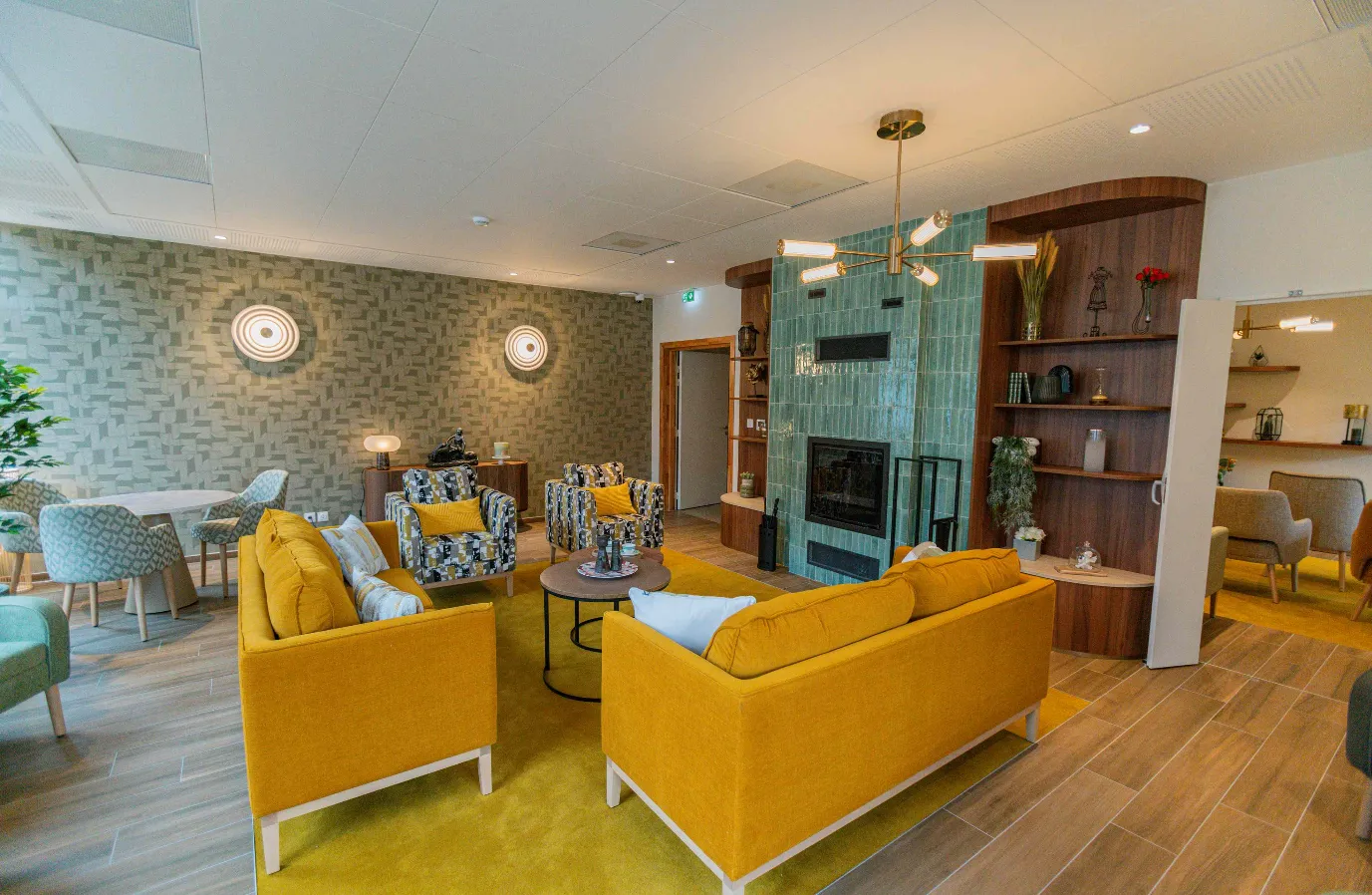 Shared lounge of the OBEO Toulouse-Guillaumet senior residence: yellow sofas, patterned armchairs and a green zellige-tiled fireplace, bathed in natural light
